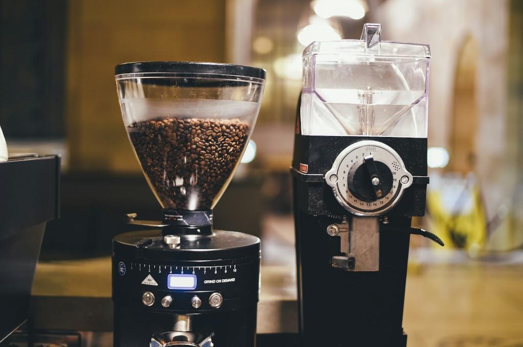 Two professional burr coffee grinders on a cafe counter — an espresso grind-on-demand grinder with beans in the hopper and a larger shop grinder with an adjustment dial